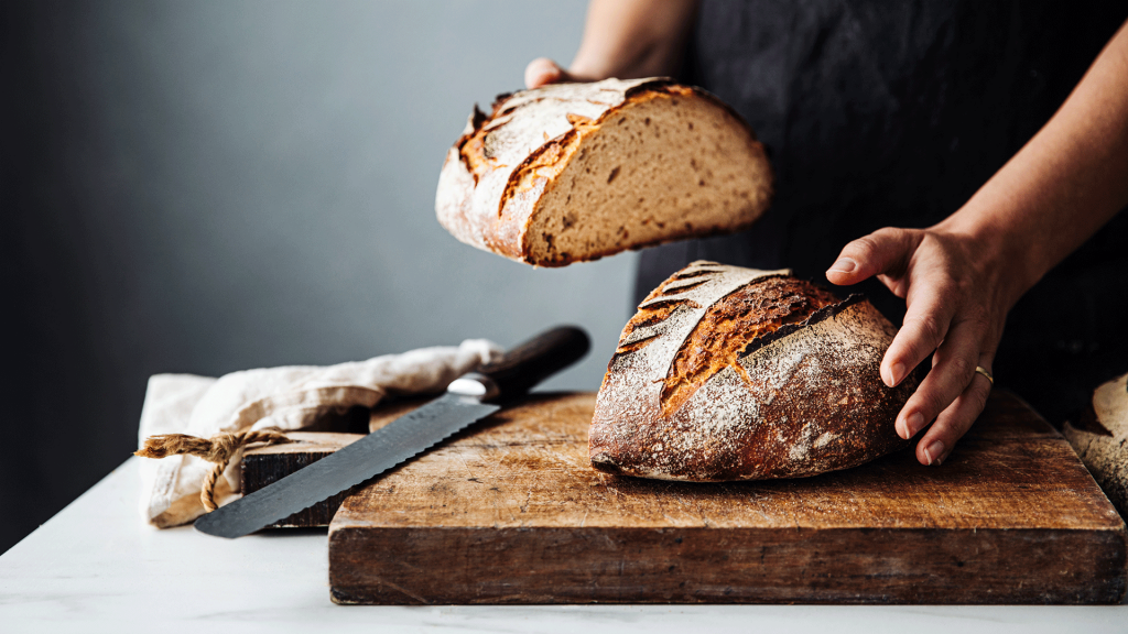 Eine Brothälfte, sowie ein Brotmesser liegen auf einem Holzbrett, während eine Hand die zweite Brothälfte hält