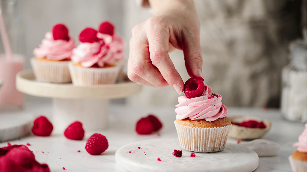 Eine Hand legt Himbeeren zur Zierde auf einen Cupcake, während weitere Cupcakes im Hintergrund zu sehen sind.