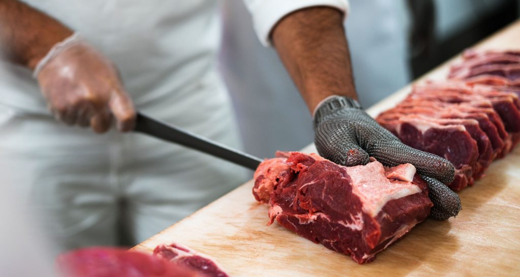 Eine Person zerteilt mit der rechten Hand ein Stück Fleisch mit einem Fleischermesser, welches sie mit der linken, mit einem Handschuh bekleideten Hand, festhält