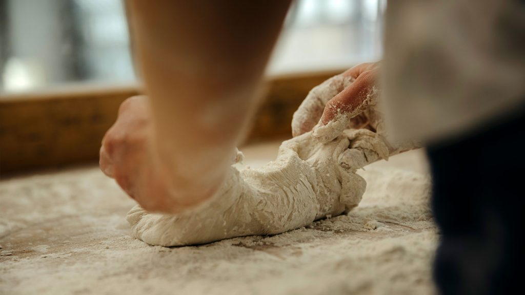 Zu sehen sind zwei Hände, welche auf einer bemehlten Arbeitsfläche Teig kneten